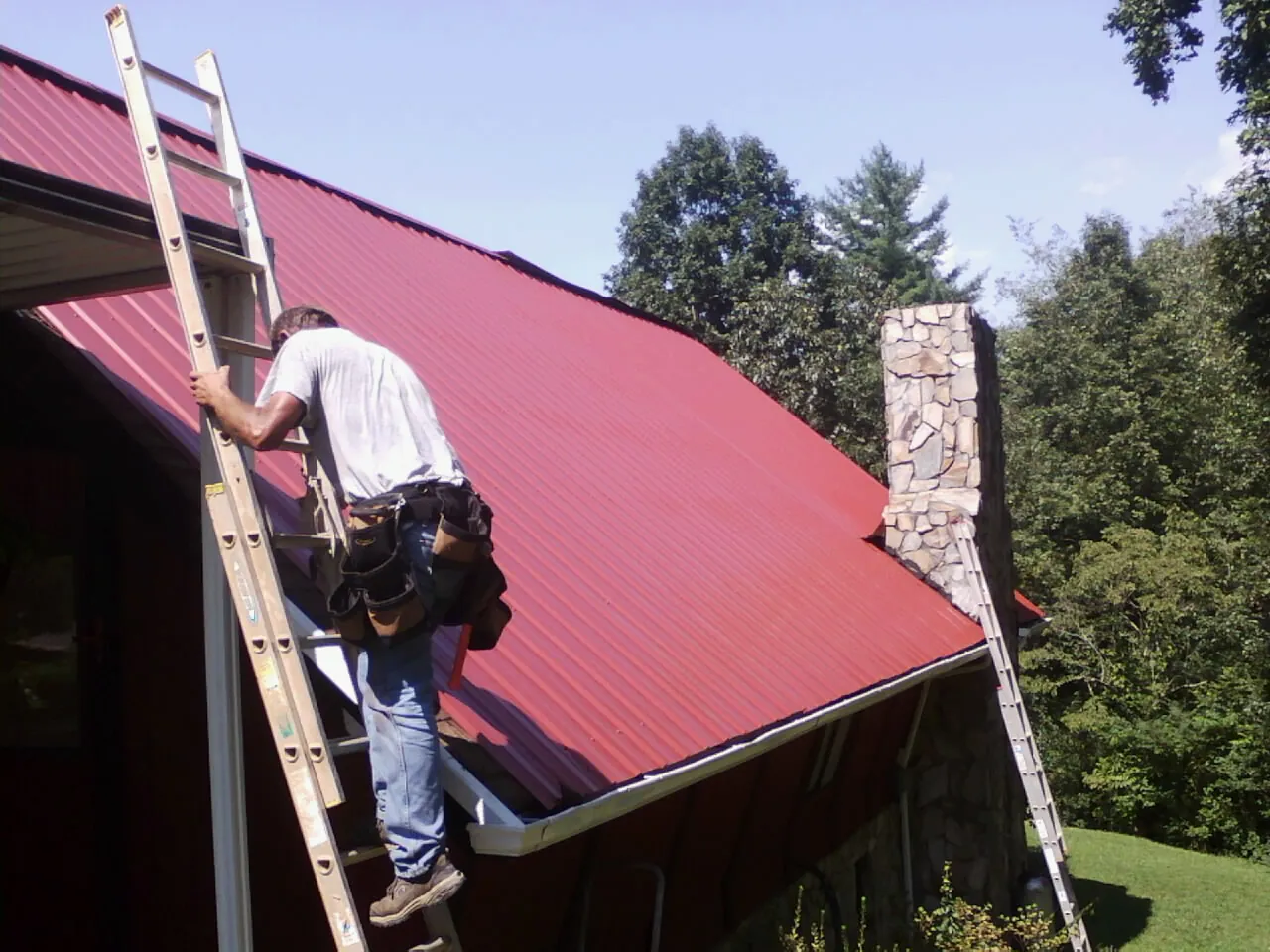 Roofer on a ladder beside a red metal roof and stone chimney — Soffit & Fascia Repair by Roofing Brothers in Yamhill